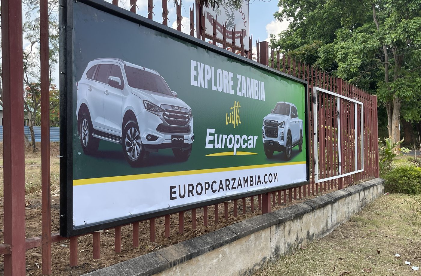 Europcar Explore Zambia billboard mounted along a roadside fence